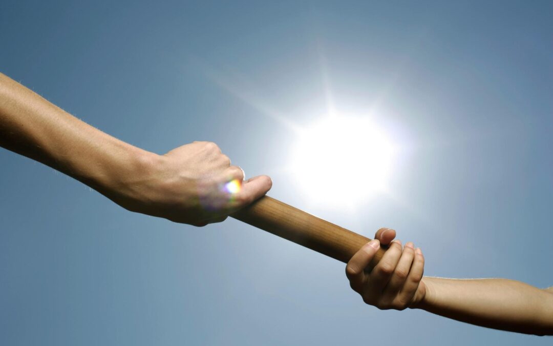Close-up of two hands passing a baton under a bright sky, symbolizing business succession and leadership transition.